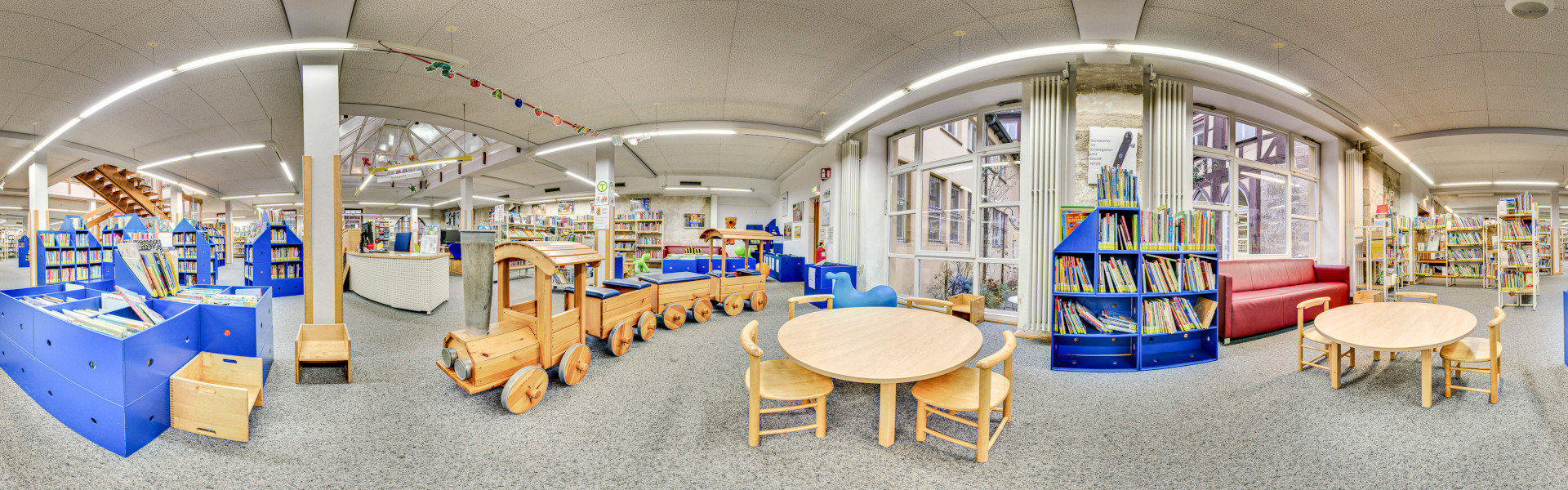 Ein Rundbild der Kinderbücherei mit großem Zug, Regalen mit Medien und Sitzgelegenheiten