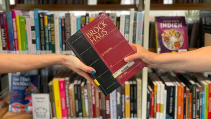 Eine Person übergibt einer anderen in einer Bibliothek einen Brockhaus-Band. Die Übergabe symbolisiert den Ausleih- oder Fernleihservice zwischen Bibliotheksnutzenden und Mitarbeitenden.